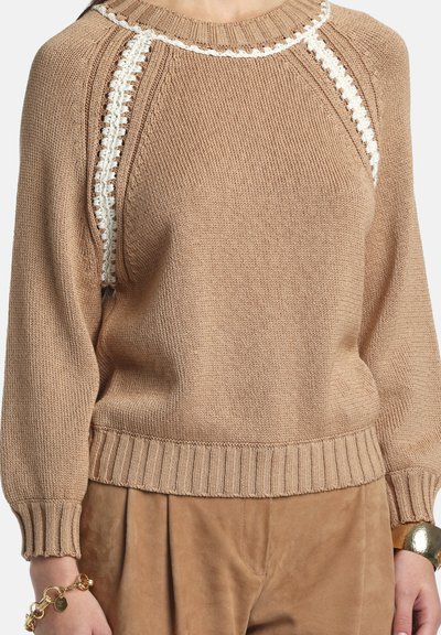 Pulover bej tricotat cu tiv zimțat, având detalii albe de-a lungul cusăturilor raglan și un decolteu rotund. Textură moale și croială lejeră.