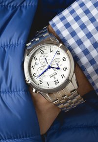 Orologio in acciaio inossidabile argento con quadrante bianco, numeri arabi neri, lancette blu e bracciale coordinato. Indossato sopra una giacca blu trapuntata.