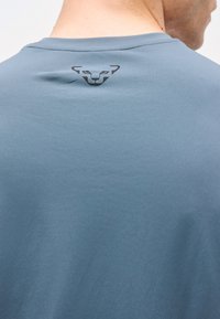 Άνδρας που φοράει ανοιχτό μπλε πουκάμισο με ένα μαύρο στιλιζαρισμένο λογότυπο ζώου στο πάνω μέρος της πλάτης κοντά στον λαιμό.