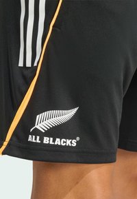 Černé sportovní šortky vyrobené z lehké tkaniny, s bílými pruhy adidas, oranžovým bočním panelem a bílým logem kapradiny s nápisem "ALL BLACKS".