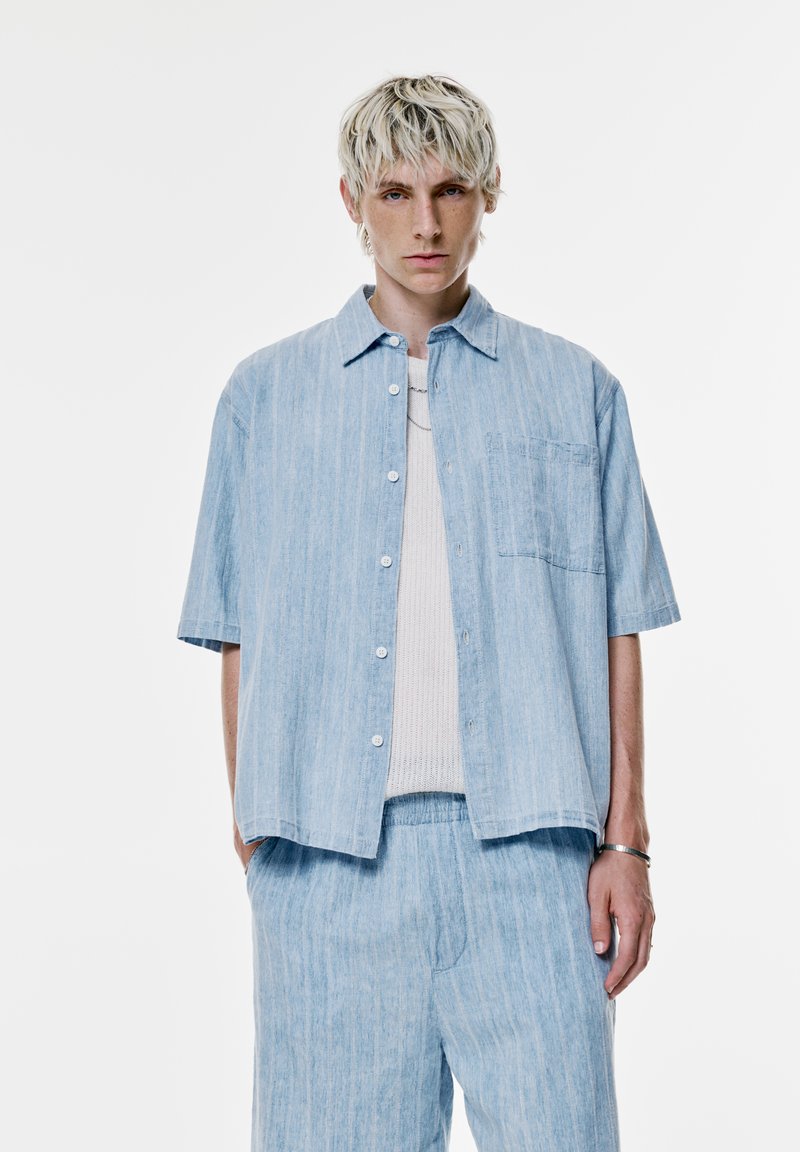 Camicia blu chiaro a maniche corte con texture a righe, bottoni frontali e taschino sul petto, abbinata a pantaloni rigati coordinati, caratterizzati da una vestibilità comoda.