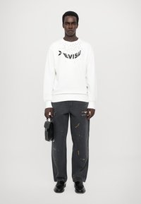 Hvid sweatshirt med trykt sort logo på brystet, parret med mørkegrå jeans med farvepletter. Modellen holder en sort taske.