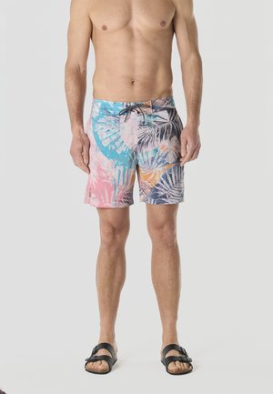 Hombre sin camiseta vistiendo pantalones cortos de baño con estampado tropical multicolor y sandalias negras, de pie contra un fondo neutro.