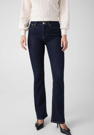 Femme portant un jean bootcut bleu foncé, un pull en maille torsadée blanc et des talons hauts noirs pointus avec un détail de boucle.