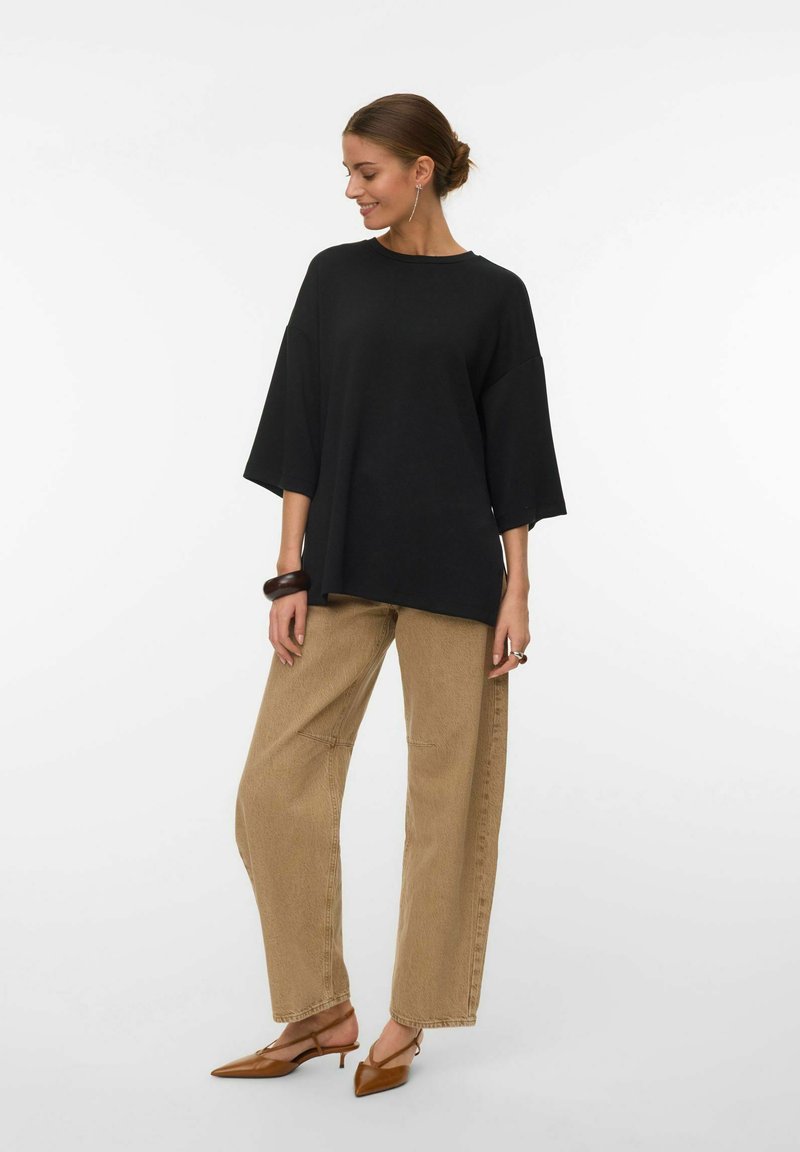 Ženska, oblečena v ohlapno črno majico s tremi četrtinami rokavi, bež široke hlače, rjave koničaste balerinke in temen zapestni trak, stoji bočno.