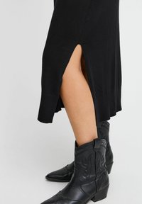 Vestido de punto negro con aberturas laterales, combinado con botas vaqueras de cuero negro que presentan detalles de costura y un tacón bajo.
