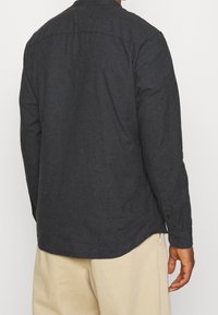 Hombre vestido con una camisa de manga larga gris oscuro y pantalones beige claros, visto desde atrás con un fondo claro y liso.