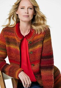 Strickjacke in Rot-, Orange- und Gelbtönen mit einem runden Kragen, fünf Knöpfen und einem strukturierten Stoff. Darunter ein rotes Oberteil getragen.