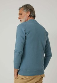 Hombre con cabello y barba canosos, vistiendo un suéter azul de manga larga y pantalones beige, girado ligeramente hacia un lado con las manos en los bolsillos.