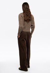 Mujer con cabello largo y ondulado, vestida con un suéter marrón suave y pantalones de pana oscuros y sueltos, con las manos en los bolsillos, de espaldas.