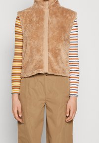 Fluffig, tan ärmlös väst med hög krage och kontrasterande beige dragkedja. Bärs över en flerfärgad, randig långärmad skjorta.