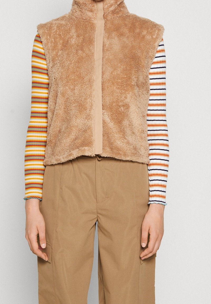 Fluffig, tan ärmlös väst med hög krage och kontrasterande beige dragkedja. Bärs över en flerfärgad, randig långärmad skjorta.