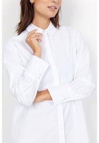 Camicia bianca con bottone realizzata in tessuto morbido. Presenta un colletto classico, maniche lunghe con polsini abbottonati e un design a taglio dritto.