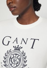 Felpa bianca con logo "GANT" blu navy e design emblema dettagliato, caratterizzata da scollo rotondo e tessuto strutturato.