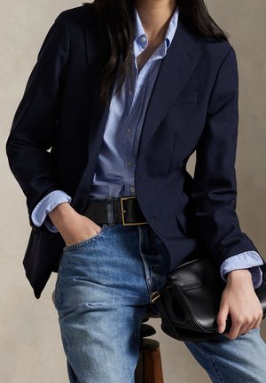 Personne portant un blazer bleu marine sur une chemise bleu clair boutonnée, un jean bleu, une ceinture noire et tenant un sac en cuir noir.