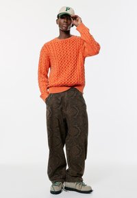 Lys orange stickad tröja med texturerat mönster, tillsammans med mörka mönstrade byxor. Modellen bär en beige keps med en grön bokstav.