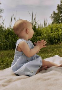 Lichtblauwe mouwloze jurk met een geraffineerde rok en kleine gele bladeren, gedragen door een zittende baby op een zacht crèmekleurig deken in het gras.