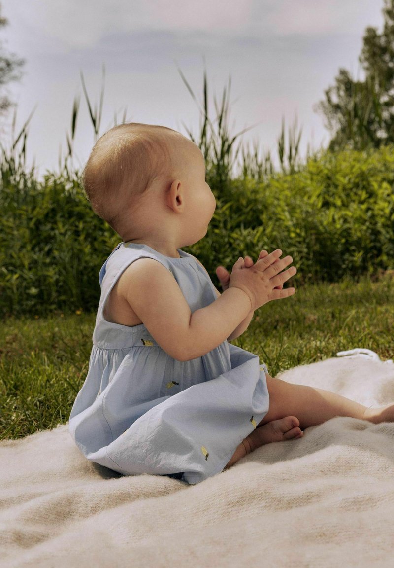 Lichtblauwe mouwloze jurk met een geraffineerde rok en kleine gele bladeren, gedragen door een zittende baby op een zacht crèmekleurig deken in het gras.
