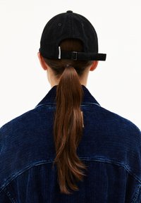 Schwarze Cordmütze mit verstellbarem Hinterband, getragen zu einer dunkelblauen Cordjacke mit strukturiertem Rippenmuster. Die Haare sind zu einem Pferdeschwanz gestylt.