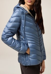 Lichtblauwe pufferjack met een glanzende textuur, horizontale stiksels, ritssluiting en een capuchon met een contrasterende crème voering.