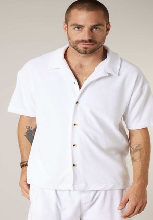 Homme aux cheveux courts et à la barbe, portant une chemise blanche à manches courtes boutonnée et un pantalon assorti, montrant des tatouages sur les deux avant-bras.