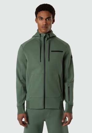 Uomo che indossa una felpa verde con cerniera, tasche frontali, coulisse e pantaloni verdi abbinati, su uno sfondo chiaro e semplice.