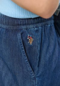 Pantaloni din denim cu talie elastică; prezintă un mic detaliu brodat pe buzunarul din față; culoare albastru mediu; textură netedă.