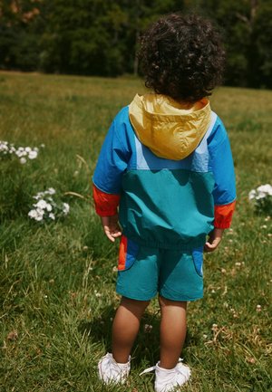 Kind mit lockigem Haar, das eine bunte Jacke und Shorts trägt, steht auf Gras und blickt nach hinten, umgeben von weißen Blumen und grünen Bäumen.