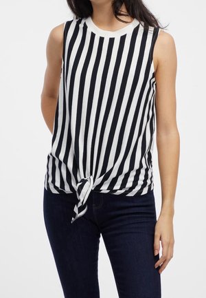 Bezrękawnik w czarno-białe pionowe paski z wiązaniem z przodu, zestawiony z ciemnoniebieskimi dżinsami na osobie na jednolitym tle.