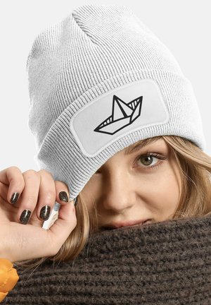 White knit beanie featuring a rectangular patch with a black paper boat design. The texture is ribbed and the fabric appears soft.