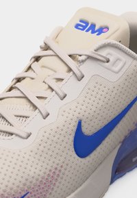 Κλείσιμο σε μπεζ αθλητικό παπούτσι με μπλε λογότυπο Nike, υφασμάτινες πλευρές, μπεζ κορδόνια και λογότυπο «AIR» σε μπλε και ροζ στην επενδυμένη γλώσσα.