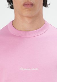T-shirt en coton rose à col rond avec col côtelé. Présente une broderie "Originals Studio" en blanc sur le devant. Texture lisse.