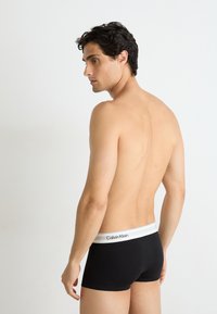 Zwarte katoenen boxershorts met een witte tailleband waarop het "Calvin Klein" logo staat, model van achteren weergegeven, staande tegen een lichte achtergrond.