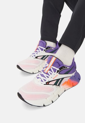 Weiße und lila Reebok-Sneaker mit schwarzen und orangefarbenen Akzenten, getragen mit weißen Socken und schwarzen Hosen auf hellem Hintergrund.