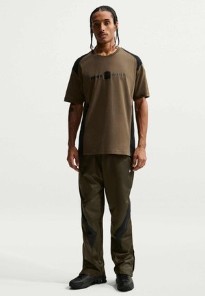 Junger Mann mit geflochtenem Haar, der ein olivgrünes und schwarzes Nike Shox T-Shirt, passende Hose und schwarze Sneakers trägt und vor einem schlichten Hintergrund steht.