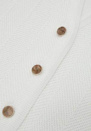 Fehér, texturált szövet cikk-cakk mintával, amelyen három kerek, márványozott barna gomb található egy átlós varrással.