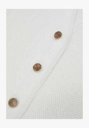 Fehér, texturált szövet cikk-cakk mintával, amelyen három kerek, márványozott barna gomb található egy átlós varrással.