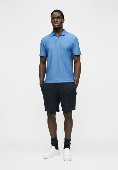 Άνδρας που φορά μπλε πόλο με κοντά μανίκια, μαύρο σορτς, σκούρες κάλτσες και λευκά αθλητικά παπούτσια, στέκεται μπροστά σε απλό λευκό φόντο.