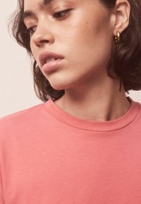 Lyserød bomulds-T-shirt med rund hals. Stoffet virker blødt og glat. Personen har kort, mørkt hår og gyldne hoop-øreringe.