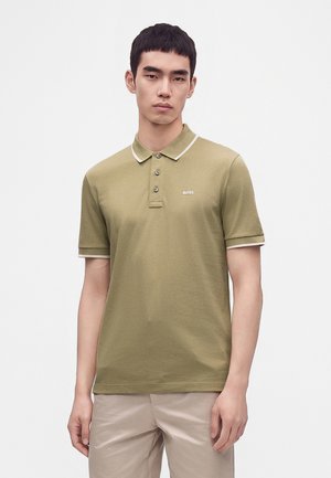 Mand iført en olivengrøn kortærmet poloshirt med hvid kant og beige bukser, stående mod en ensfarvet lys baggrund.