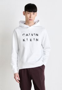Bela kapuca s potiskom "CALVIN KLEIN" v črni barvi. Ima kenguru žep in kapuco s vrvico. Združeno z rdečimi hlačami.
