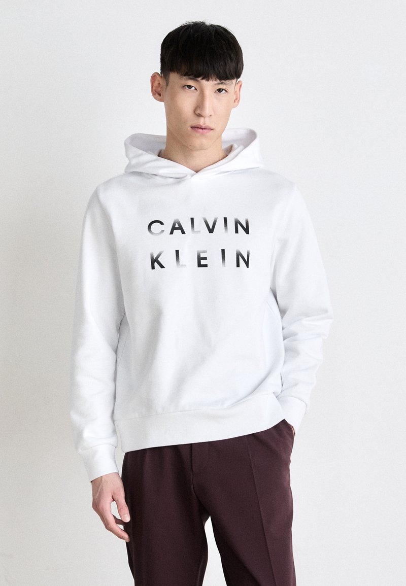 Bela kapuca s potiskom "CALVIN KLEIN" v črni barvi. Ima kenguru žep in kapuco s vrvico. Združeno z rdečimi hlačami.