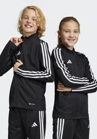Zwarte sportjacks met witte drie-streep accenten, hoge kragen, ritsvoorkanten en een textuurstof met het "AEROREADY" logo aan de onderkant.