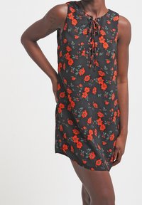 Ärmelloses schwarzes Kleid mit rotem Blumenmuster, mit gebundenem Ausschnitt, präsentiert von einer Person mit einer Hand auf der Hüfte und der anderen entspannt.