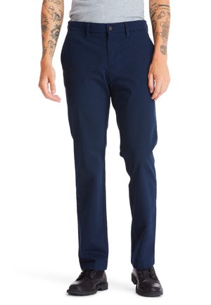 Navyblauwe katoenen broek met een rechte snit, voorzien van een knoopsluiting en voorzakken, gedragen met zwarte veterschoenen.