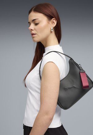 Donna con capelli rossi indossa una maglietta bianca, porta una borsa a tracolla nera Puma con etichetta bordeaux, rivolta verso sinistra su sfondo grigio.