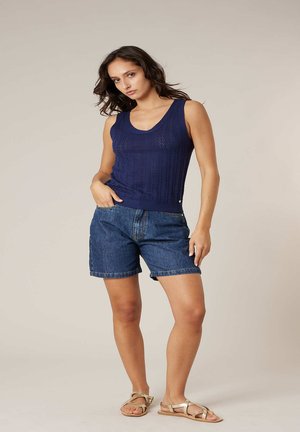 Femme debout, une main dans la poche de son short en denim, portant un haut sans manches en maille bleu marine et des sandales plates à lanières couleur beige.