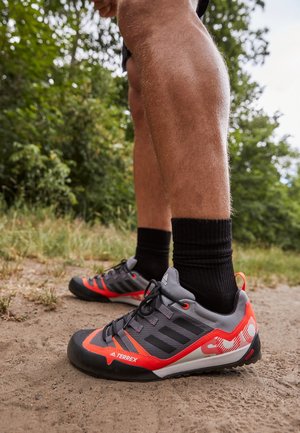 Beine mit schwarzen Socken und grauen und roten Adidas Terrex Wanderschuhen stehen auf einem Erdweg mit grünem Laub im Hintergrund.
