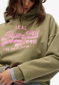 Hoodie verde oliva con testo in strass rosa "REAL Superdry" e caratteri giapponesi. Le maniche presentano un dettaglio di tessuto a righe sotto.