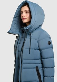 Plava jakna s kapuljačom, punjena, s visokim ovratnikom, prilagodljivim šnurbanjem i bočnim džepovima na patentni zatvarač; sadrži okruglu oznaku na rukavu.